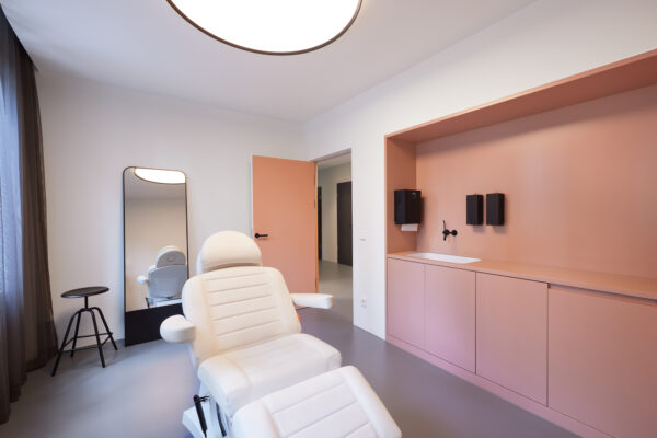 Das Behandlungszimmer beinhaltet gleiche Farbelemente (Hellblau, Pastell orange) mit einem Behandlungsstuhl, Spiegel, Waschbecken und Schränken.