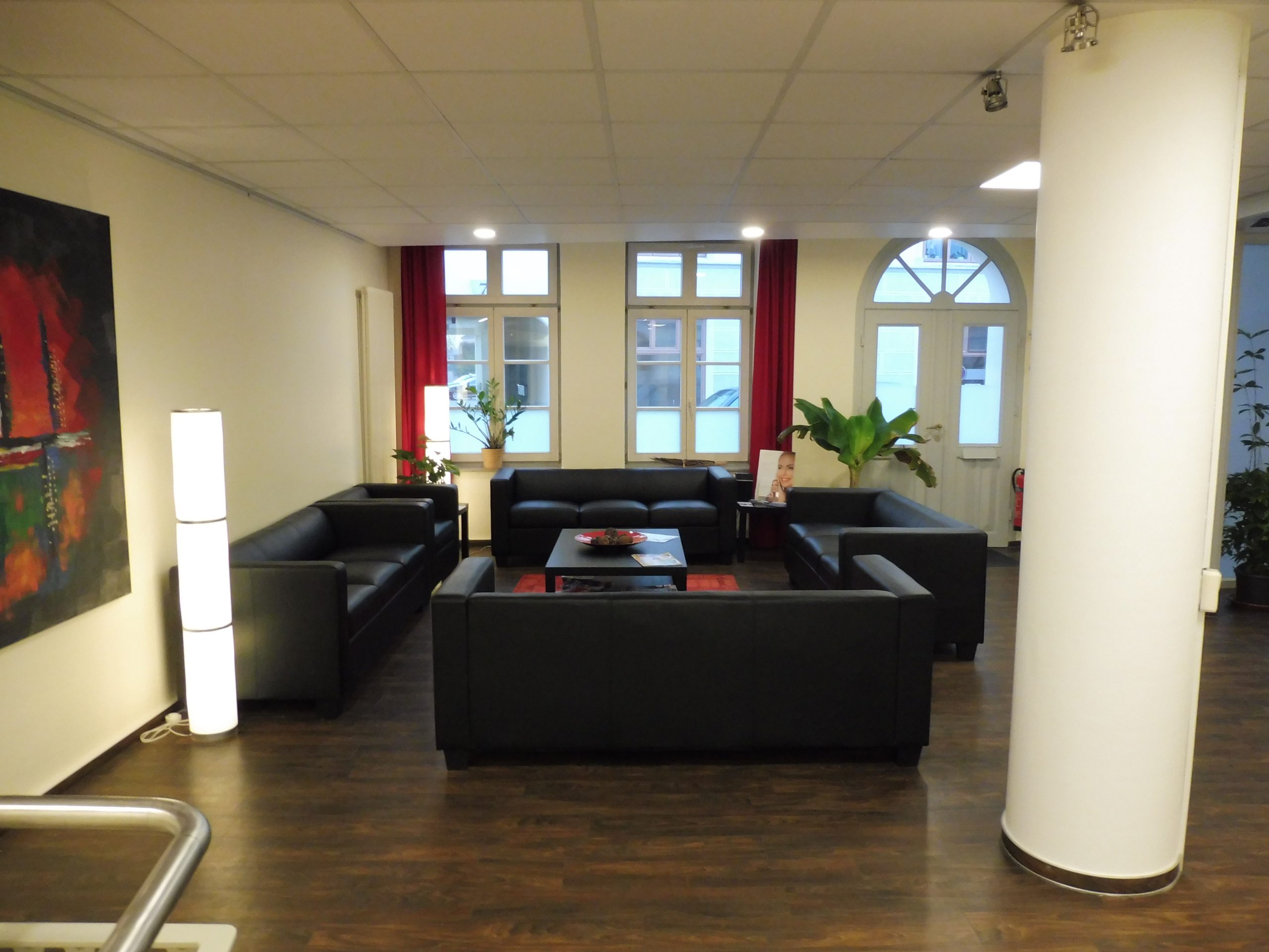 Der Wartebereich hat vier schwarze Sofagruppen um einem schwarzen Beistelltisch platziert. Vorhänge und Poster sind ebenfalls in einem schwarz-roten Frabschema. Pflanzen und viele Lichter runden den Raum ab.