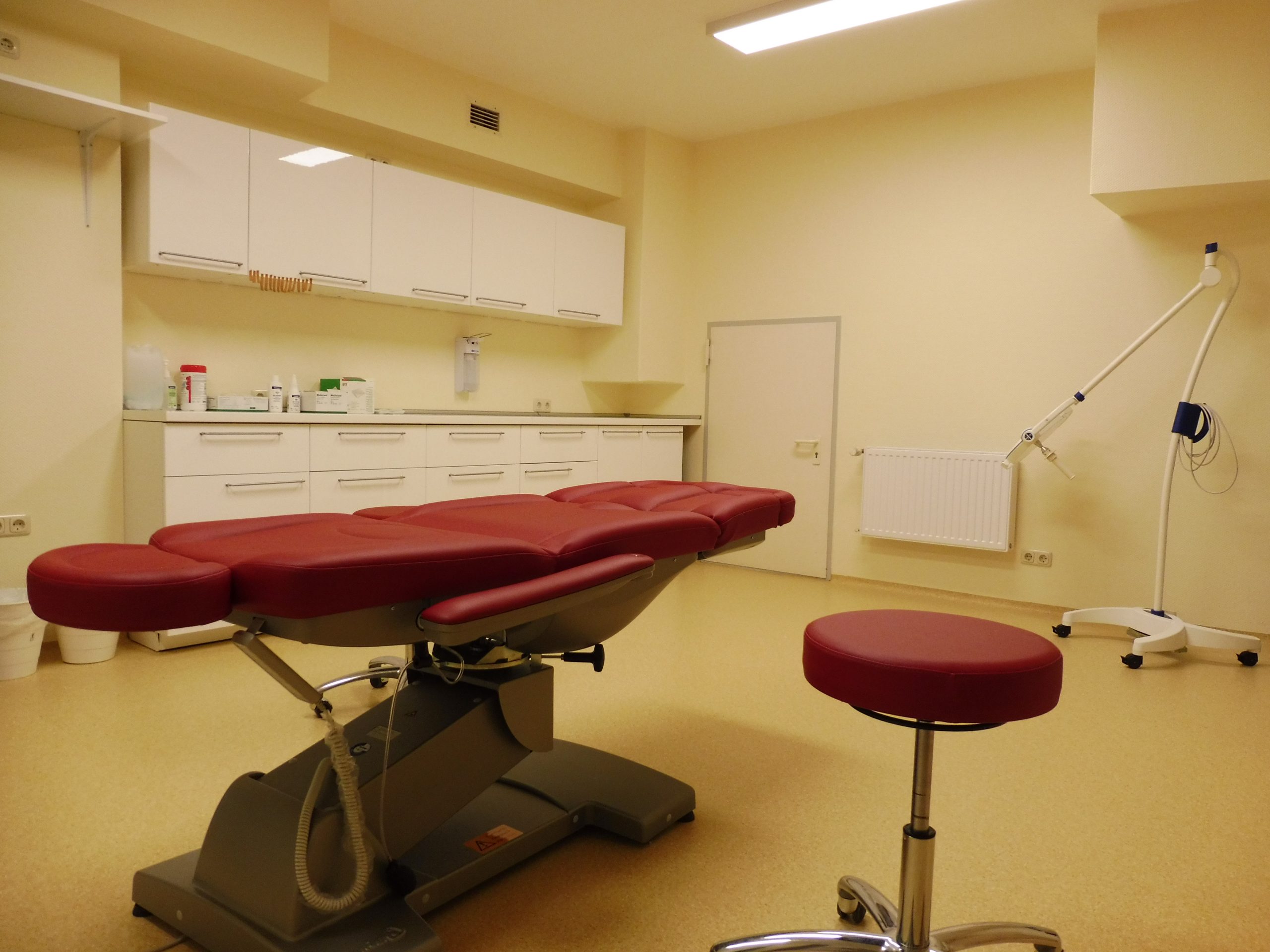 Das Behandlungszimmer hat einen roten Behandlungsstuhl sowie einen roten Rollhocker. An der Wand befinden sich Ober- und Unterschränke. Diverse technische Geräte sind ebenfalls zu finden.