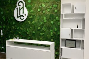 Der Empfang ist mit weißen Möbeln ausgestattet. Die Wand dahinter ist aus grünem Kunststoffrasen. Mitte ist ein Logo 