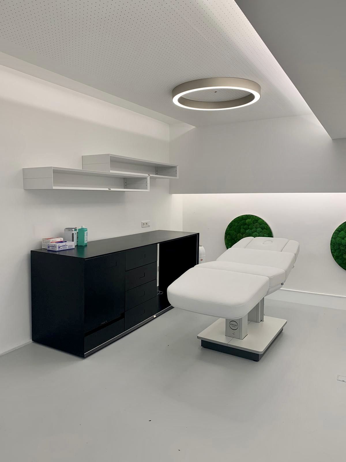 Das Beratungszimmer ist im gleichen Stil gestaltet. Der weiße Patientenstuhl steht neben den Regalen und Schränken, die Materialien für die Behandlung verstauen. An der Wand sind runde Kunststoffrasen dekorativ befestigt.