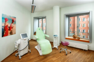 Ein weiteres Behandlungszimmer ausgestattet mit einem Patientenstuhl, hochwertigem Equipment für andere Behandlungszwecke. Außerdem ist es ebenfalls dekorativ eingerichtet mit einem Poster und durch mehrere Fenster hell erleuchtet.