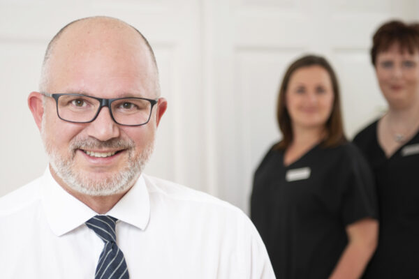 Prof. Dr. Lars Steinsträßer steht lächelnd im Fokus vor der Kamera. Zwei seiner Mitarbeiterinnen stehen im Hintergrund lächelnd.