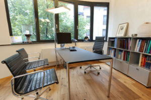 Büro von Dr. med. Jörg Blesse mit Computertisch und -Stuhl und Ordnerkabinett dahinter. Gegenüber zwei Stühle für Gäste/Patienten. Das Büro hat eine große Fensterwand.