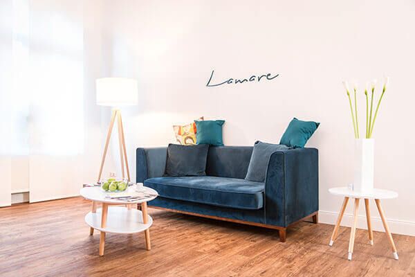 Der Wartebereich ist hell eingerichtet mit einer blauen Couch und zwei Beistelltischen mit Info-Materialien und Deko. An der Wand ziert sich die Schrift 