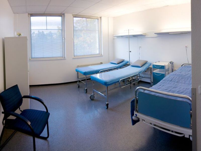 Das Patientenzimmer zeigt zwei Patientenliegen, ein Krankenbett, ein Schrank und Stuhl sowie Regale.