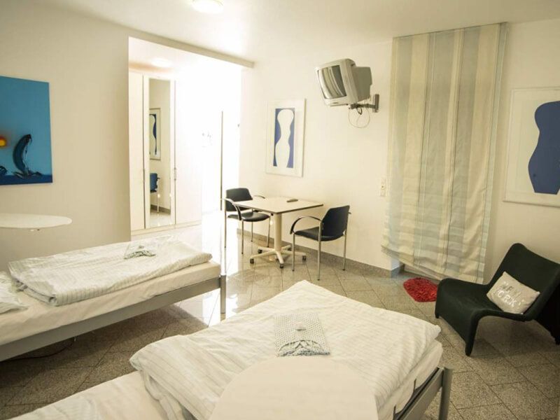 Das Patientenzimmer hat zwei Krankenbetten, Fernseher und ein Tisch mit zwei Stühlen. An den Wänden hängen gerahmte Poster.