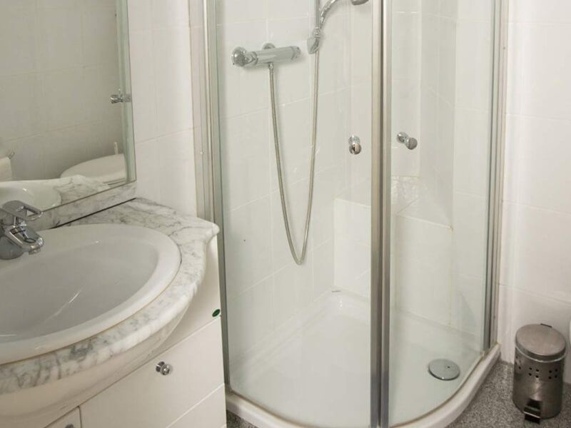 Das Badezimmer für den Patienten ist in einer weißen Ausstattung und umfasst Waschbecken mit Unterschrank, darüber ein Spiegel und rechts davon die Dusche und Mülleimer.