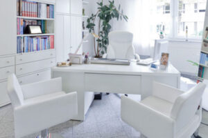 Das Büro von Dr. Manuel Hrabowski. Im Vordergrund ist sein PC-Schreibtisch mit zwei gegenüber stehenden Stühlen für Patienten. Schränke mit Büchern und ein Spiegel stehen jeweils links und rechts vom Tisch. Die Möbel sind fast ausschließlich in weiß gehalten.