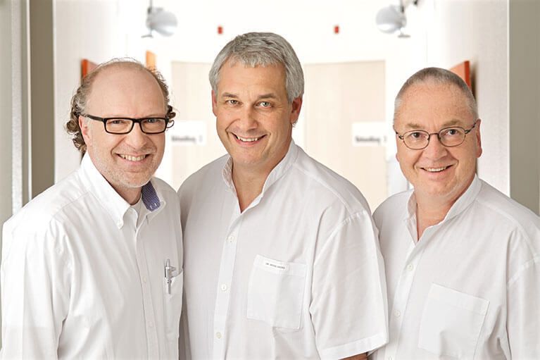Michael Schneider und zwei seiner Kollegen lächeln in die Kamera. Alle drei stehen im Gang in der Praxis.