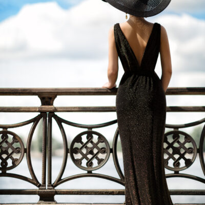 Woman Back Side View in Hat and Evening Dress Outdoor. Fashion Model Rear View looking away. Luxury Elegant Lady in long Gown at Promenade Fence looking at Sky