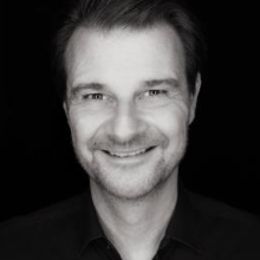 Headshot von Dr. med. Claudius Kässmann lächelnd in schwarz-weiß. Aufgenommen vor einem dunklen Hintergrund.