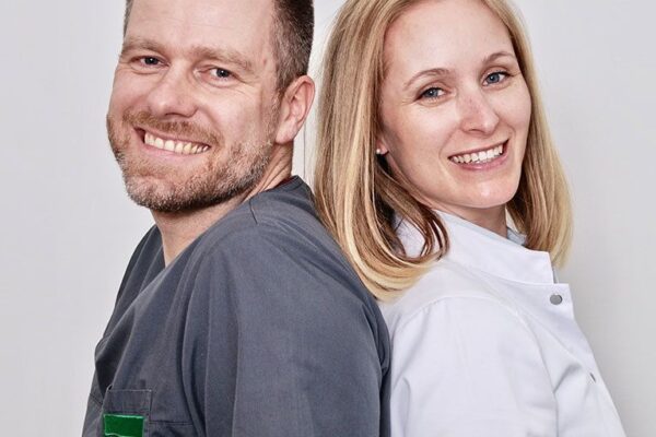 Dr. med. Juliane Finke und Dr. med. Stephan Frantzen stehen mit ihren Arztkitteln Rücken and Rücken und lächeln in die Kamera.