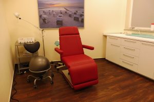 Das Behandlungszimmer ist ausgestattet mit einem roten Behandlungsstuhl, einem schwarzen Rollhocker, Unterschränken, technischen Geräten und einem Poster an der Wand.