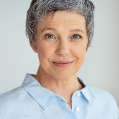 Closeup face of senior business woman standing against grey background with copy space. Portrait of successful woman in blue shirt feeling confident and looking at camera. Happy mature woman face standing.