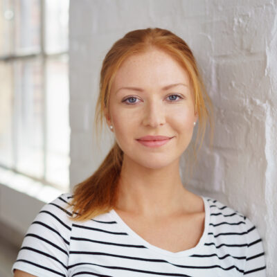 Thoughtful pretty young redhead woman in a striped t-shirt leaning on a white painted brick interior wall looking at the camera with a quiet smile
