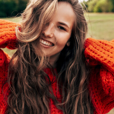 Outdoor portrait of a smiling young woman wearing a knitted orange sweater posing on nature background. The beautiful female has a joyful expression, resting in the park.