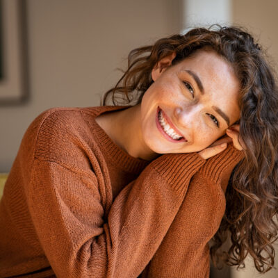 Happy young woman sitting on sofa at home and looking at camera. Portrait of comfortable woman in winter clothes relaxing on armchair. Portrait of beautiful girl smiling and relaxing during autumn.