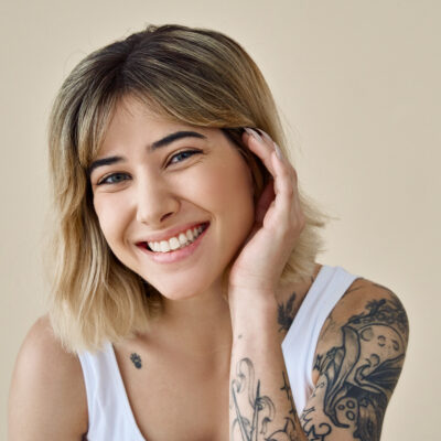 Smiling blond pretty happy girl beauty female model with short blonde hair and tattoos looking at camera touching face pampering advertising skin care isolated at beige background. Portrait.