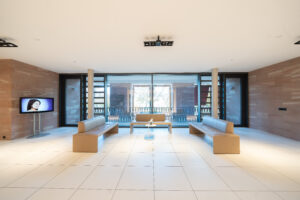 Modernes und einladens Foyer der Schönheitsklinik mit hellen Fliesenböden, beigen Ledersofas, einem großen Fenster mit Blick ins Grüne und einem Bildschirm, der ästhetische Behandlungen präsentiert. Die Atmosphäre strahlt Ruhe und Eleganz aus.