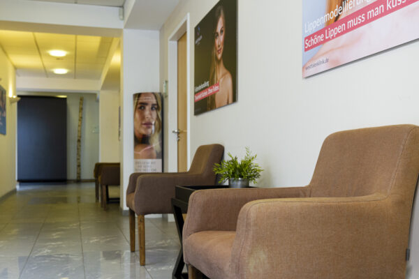 Das Bild zeigt einen Wartezimmerbereich in der Westfalenklinik. Im Vordergrund stehen zwei braune Sessel mit einem kleinen Tisch dazwischen, auf dem sich eine Topfpflanze befindet. An den Wänden hängen Werbeposter. Im Hintergrund ist ein Korridor zu sehen, der weitere Wartezimmermöbel zeigt. Das Gesamtbild vermittelt einen sauberen, modernen und einladenden Eindruck, der typisch für eine Schönheitsklinik ist.