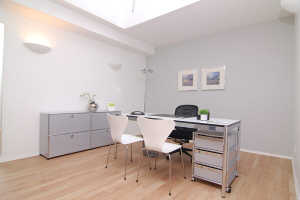 Das Bild zeigt ein modern eingerichtetes Büro in der Westfalenklinik. Es ist hell und freundlich gestaltet mit weißen und hellgrauen Wänden, hellem Holzboden und modernen, silbernen Metallmöbeln. Ein Schreibtisch mit zwei weißen Stühlen steht im Mittelpunkt, daneben ein grauer Schrank und ein Rollcontainer. An der Wand hängen zwei Bilder mit Landschaftsmotiven. Die Atmosphäre wirkt ruhig und professionell.