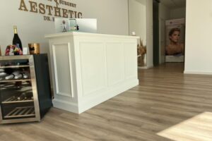 Heller und einladender Empfangsbereich einer Schönheitsklinik. Im Vordergrund steht ein weißer Empfangstresen mit dekorativen Elementen. An der Wand ist der Name der Klinik angebracht. Links vom Tresen befindet sich ein Weinkühlschrank mit diversen Flaschen. Im Hintergrund führt ein Gang zu weiteren Behandlungsräumen. Ein Werbebanner für Schönheitsbehandlungen ist sichtbar. Der Boden ist mit hellem Holzparkett ausgelegt.